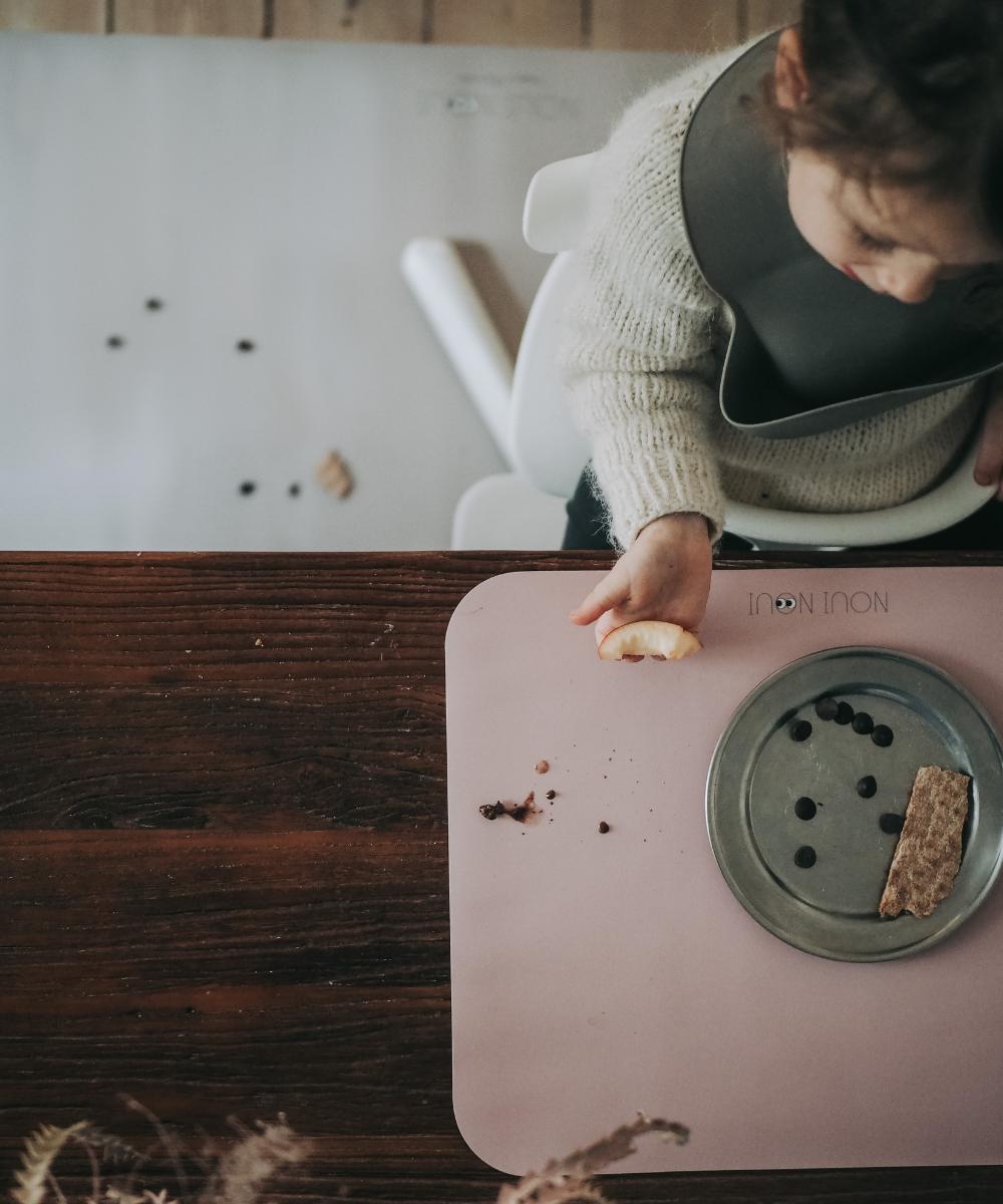 Barn med Noui Noui hagesmæk Warm Gray ved bordet med rosa dækkeserviet